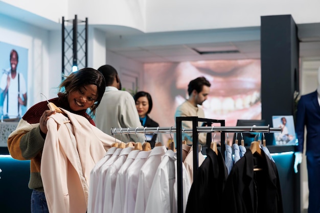 Afroamerikanische Frau hält ihr Outfit am Körper und überprüft Größe und Stil, bevor sie in einer Boutique einkauft. Kunde untersucht formelles Hemd, das auf einem Hänger in einem Bekleidungsgeschäft passt