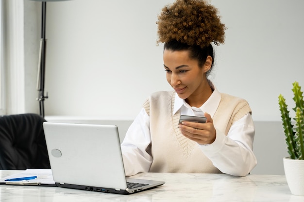 Afroamerikanische Frau mit einer Kreditkarte an einem Tisch sitzend mit einem Laptop beim Online-Shopping