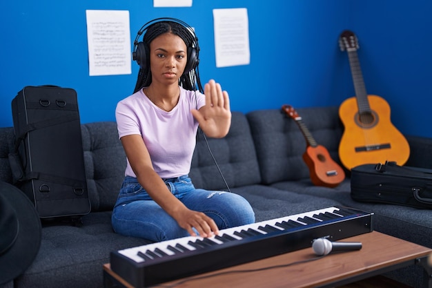 Afroamerikanische Frau mit Zöpfen spielt Klavier in einem Musikstudio mit offener Hand und macht ein Stoppschild mit ernsthafter und zuversichtlicher Ausdrucksverteidigung.