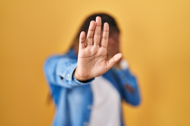 Afroamerikanische Frau mit Zöpfen steht vor gelbem Hintergrund und bedeckt die Augen mit den Händen und macht Stopp-Geste mit traurigem und ängstlichem Ausdruck, verlegenem und negativem Konzept