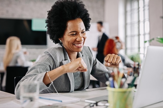 Afroamerikanische Geschäftsfrau fühlt sich zufrieden und feiert, während sie gute Nachrichten auf einem Computer im Büro liest