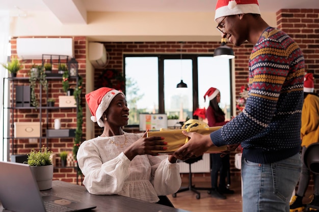 Afroamerikanische kollegen tauschen weihnachtsgeschenkbox im festlichen büro mit weihnachtsbaum aus, um winterferien zu feiern. Menschen, die saisonale Geschenke geben und sich über den Weihnachtsmann freuen.