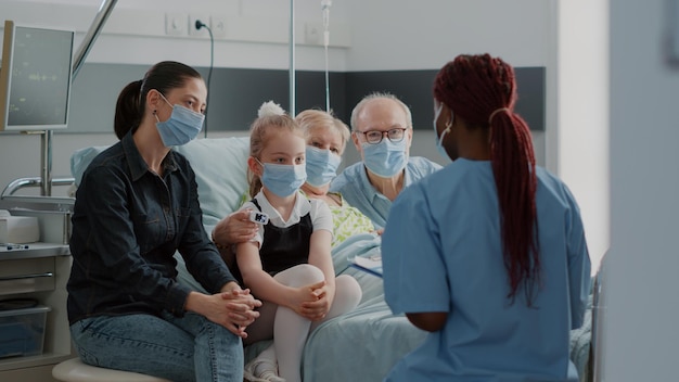 Afroamerikanische Krankenschwester, die älteren Patienten in der Krankenstation hilft, während Familienbesuche während der Covid-19-Pandemie besucht werden. Gesundheitsspezialist spricht mit alter Frau und Besuchern über Genesung.