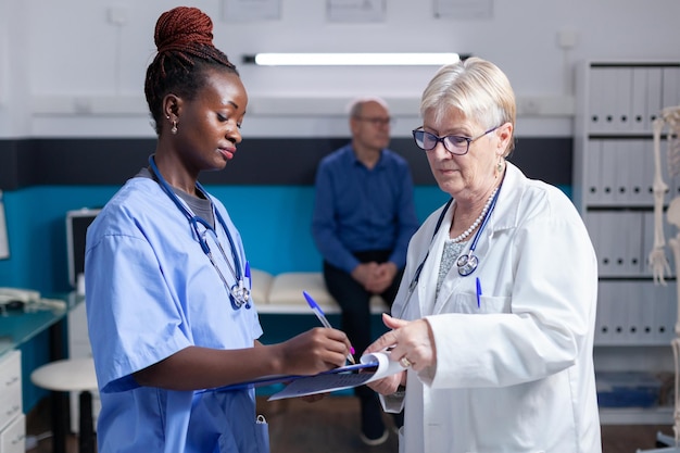 Afroamerikanische Krankenschwester, die Checkup-Papiere unterschreibt, während der Arzt die Zwischenablage im Schrank hält. Arzthelferin und Arzt, die beim Kontrollbesuch Gesundheitsinformationen und Daten in Akten einsehen