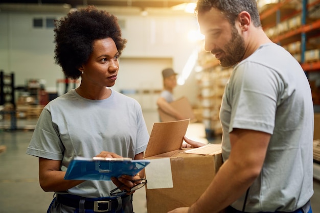 Afroamerikanische Lagerarbeiterin, die mit ihrer Kollegin spricht, während sie an einem industriellen Lagerabteil arbeitet