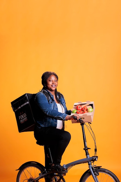 Afroamerikanische Lebensmittellieferantin fährt Fahrrad, während sie dem Kunden eine Kiste voller Bio-Gemüse für ein gesundes Mittagessen liefert, während sie das Fahrrad als Transportmittel nutzt. Essensservice und Konzept zum Mitnehmen