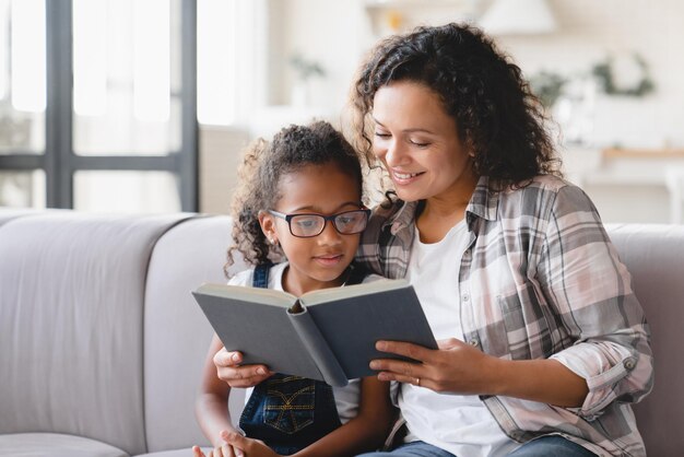 Afroamerikanische Mutter: Mutter liest Märchen, Buch, Roman mit Tochter, Mädchen lernt zusammen, hilft bei den Hausaufgaben für die Schule, Lehrerin unterrichtet Schulmädchen zu Hause, Homeschooling