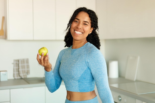 Afroamerikanische sportliche Dame hält Apfel und lächelt in die Kamera und isst frisches Obst nach dem Training in der Küche