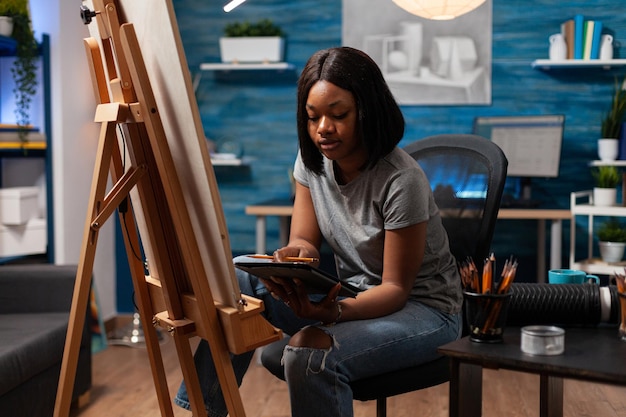 Afroamerikanische Studenten, die beim Surfen Inspiration auf dem Tablet-Computer ziehen, der vorne in einer Leinwandzeichnungsskizze sitzt, mit grafischem Bleistift im Kunstatelier. Junger Künstler, der an einem authentischen Meisterwerk der Kunst arbeitet