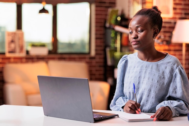 Afroamerikanische Studentin der entfernten Schule, die Notizen im Notizbuch macht und zu Hause mit Laptop lernt. Tutorials für Internetkurse, Online-Bildung und modernes E-Learning-Konzept