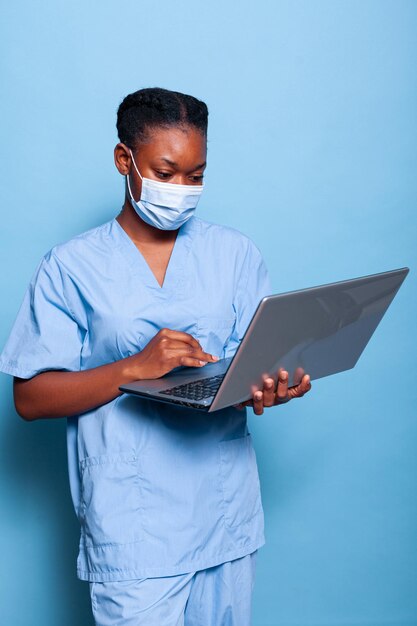 Afroamerikanische Therapeutenkrankenschwester mit schützender Gesichtsmaske zur Verhinderung einer Infektion mit Coronavirus, die Gesundheitsbehandlung auf Laptop-Computern im Studio mit blauem Hintergrund eingibt. Medizinservice