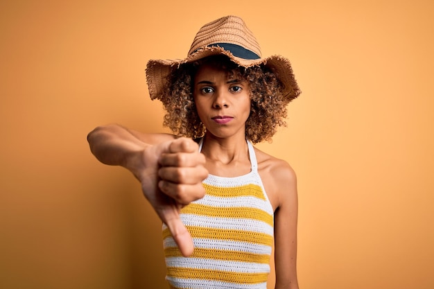 Afroamerikanische Touristin mit lockigem Haar im Urlaub, trägt Sommerhut und gestreiftes T-Shirt und sieht unglücklich und wütend aus, zeigt Ablehnung und Negativität mit der Geste des Daumens nach unten. Schlechter Gesichtsausdruck