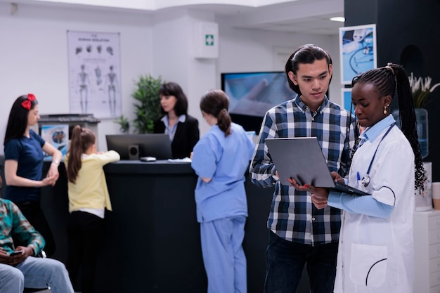 Afroamerikanischer Arzt mit Laptop und Termin im Gespräch mit asiatischem Patienten im geschäftigen Wartezimmer einer Privatklinik. Sanitäter, der sich mit einem jungen Mann unterhält, der die Laborergebnisse überprüft.