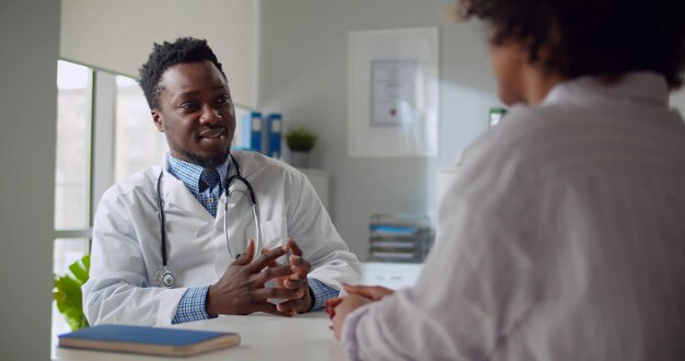 Afroamerikanischer Arzt spricht mit Patient, der am Schreibtisch im Büro sitzt