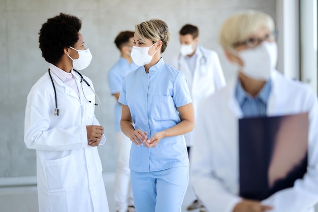 Afroamerikanischer Arzt und Krankenschwester tragen Gesichtsmasken, während sie in einem Flur der Klinik sprechen