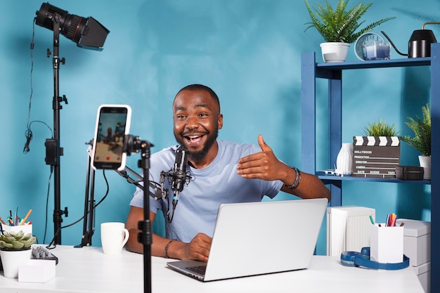 Afroamerikanischer Blogger spricht und benutzt Laptop, während er Videos auf dem Smartphone für einen Internetkanal aufnimmt. Online-Streamer überträgt Live-Übertragung per Mobiltelefon auf Stativ