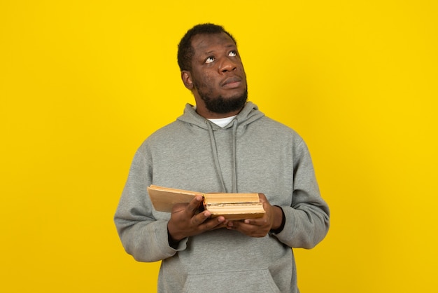 Afroamerikanischer denkender Mann, der interessante Bücher in seiner Hand liest, steht über gelber Wand.