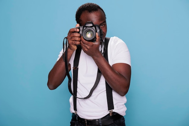 Afroamerikanischer Fotografie-Enthusiast, der ein Foto macht, während er auf blauem Hintergrund steht. Selbstbewusster professioneller Fotograf mit DSLR-Kamera beim Fotografieren. Studioaufnahme.