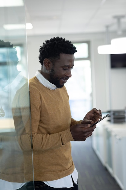 Afroamerikanischer Geschäftsmann arbeitet in einem modernen Büro, lehnt sich mit dem Smartphone an die Glaswand und lächelt. Soziale Distanzierung am Arbeitsplatz während der Coronavirus-Covid-19-Pandemie.