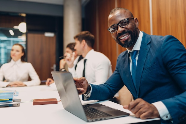 Afroamerikanischer Geschäftsmann mit schwarzem Mann, der am Tisch mit Laptop auf dem Hintergrund der Mitarbeiter des Büroteams sitzt
