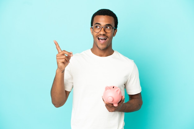 Afroamerikanischer gutaussehender Mann, der ein Sparschwein über isoliertem blauem Hintergrund hält, der die Lösung realisieren möchte, während er einen Finger hochhebt
