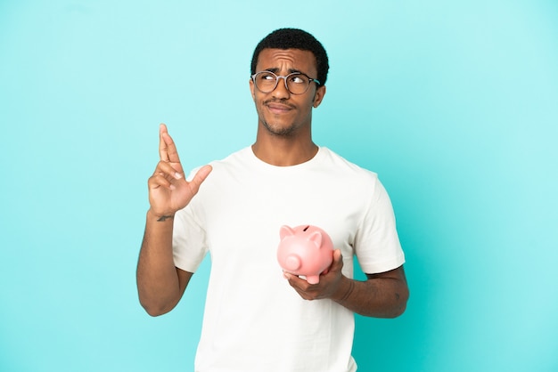 Afroamerikanischer gutaussehender Mann, der ein Sparschwein über isoliertem blauem Hintergrund hält, die Finger kreuzen und das Beste wünschen