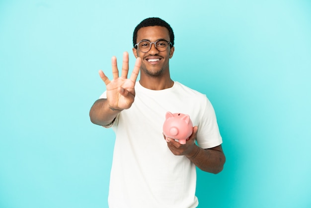 Afroamerikanischer gutaussehender Mann, der ein Sparschwein über isoliertem blauem Hintergrund hält, glücklich und zählt vier mit den Fingern