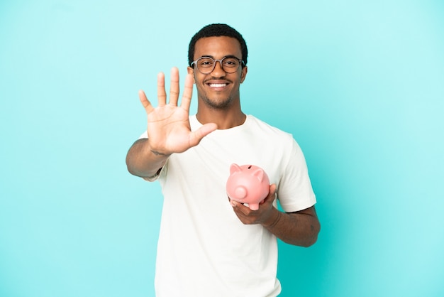 Afroamerikanischer gutaussehender Mann, der ein Sparschwein über isoliertem blauem Hintergrund hält und fünf mit den Fingern zählt