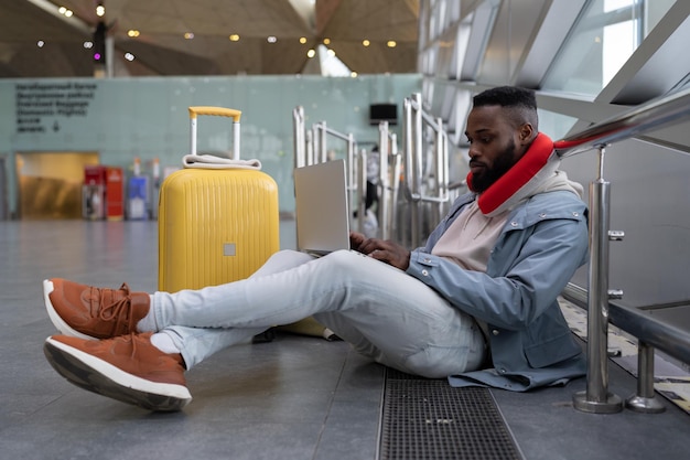 Afroamerikanischer Hipster-Typ, der am Computer auf Knien arbeitet und auf dem Boden in der Nähe des Fensters im Flughafen sitzt