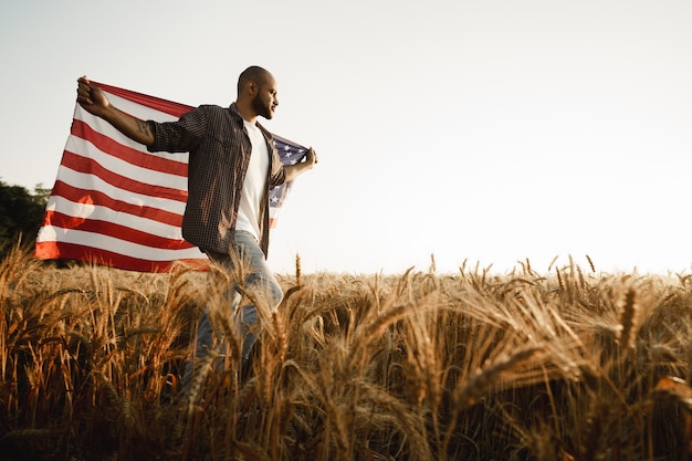 Afroamerikanischer junger Mann, der die Nationalflagge der USA durch Weizenfeld hält