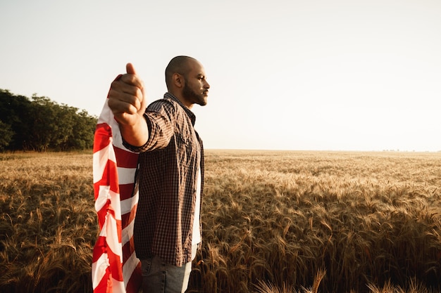 Afroamerikanischer junger Mann, der die Nationalflagge der USA durch Weizenfeld hält