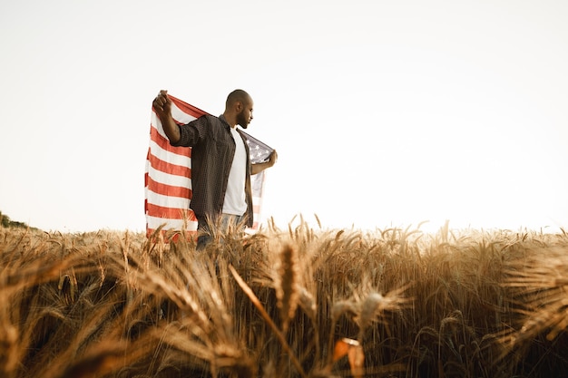 Afroamerikanischer junger Mann, der die Nationalflagge der USA durch Weizenfeld hält