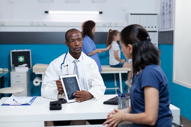 Afroamerikanischer Kinderarzt, der Tablet-Computer mit Kinderradiographie auf dem Bildschirm hält und während der Beratung im Krankenhausbüro die Behandlung der Gesundheitsversorgung mit der Mutter des Patienten bespricht. Medizinservice