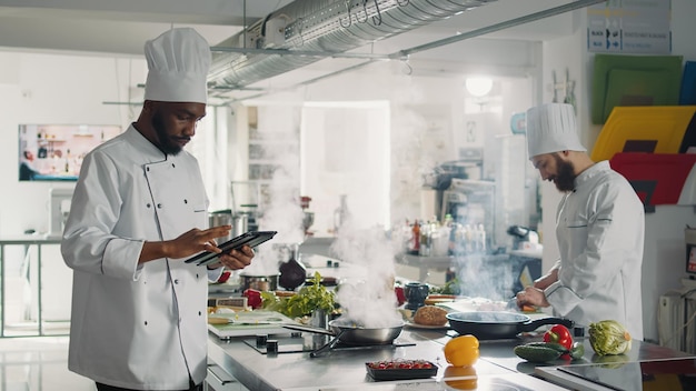 Afroamerikanischer Koch sucht Gourmet-Rezepte auf einem digitalen Tablet und verwendet ein Gerät mit Technologie, um ein professionelles Gericht zuzubereiten. Männlicher Koch in Uniform kocht gastronomische Mahlzeit von der Website.