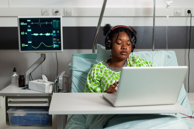 Afroamerikanischer kranker Patient, der im Bett liegt und Online-Comedy-Cartoons auf Laptop-Computer ansieht, während er sich nach einer medizinischen Operation während eines klinischen Termins in der Krankenstation erholt. Medizinservice