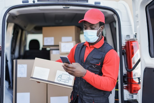 Afroamerikanischer Lieferbote mit Smartphone, der Paket hält, während er eine Sicherheitsgesichtsmaske für den Ausbruch des Coronavirus trägt