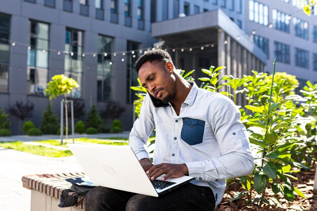 Afroamerikanischer männlicher Freiberufler, der aus der Ferne Online-Broker mit Börsenaktien arbeitet, die über Mobiltelefon und Laptop sprechen Händlerverkäufer. Mann im Stadtpark ein moderner städtischer Hintergrundgeschäftsmann der Bank