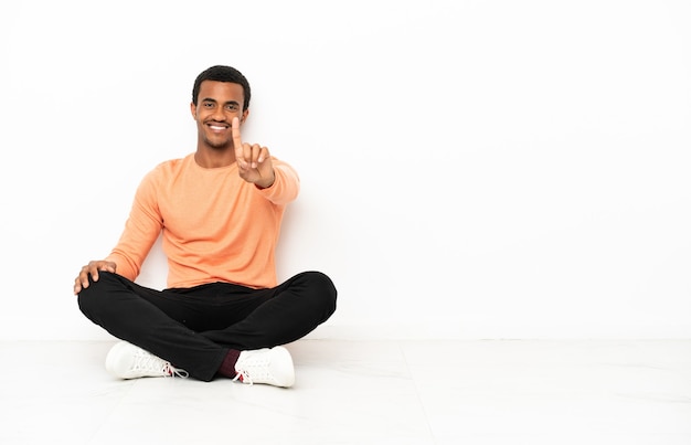 Afroamerikanischer Mann, der auf dem Boden über isoliertem Hintergrund sitzt und einen Finger zeigt und hebt