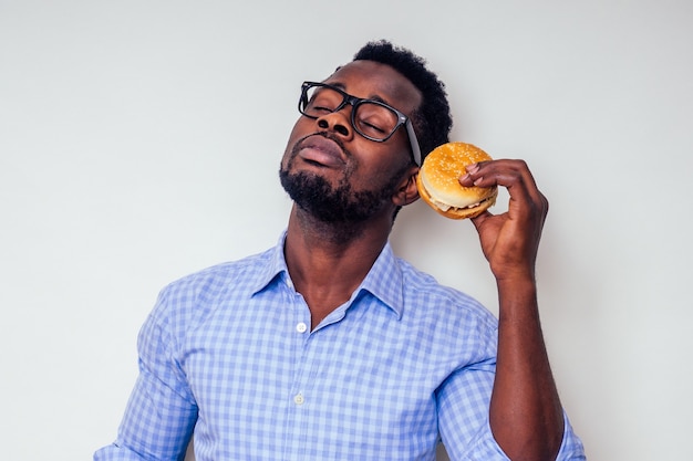 Afroamerikanischer Mann, der den Geschmack von Hamburger genießt. Gutaussehender und junger Afro-Mann in einem stilvollen Hemd und einer Brille, die einen Burger auf weißem Hintergrund hält