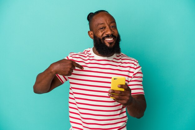 Afroamerikanischer Mann, der ein Mobiltelefon einzeln auf blauem Hintergrund hält, der mit der Hand auf einen Hemdkopierraum zeigt, stolz und selbstbewusst