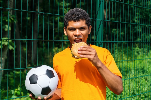 Afroamerikanischer Mann, der Fußball hält, während er gegen Zaun steht