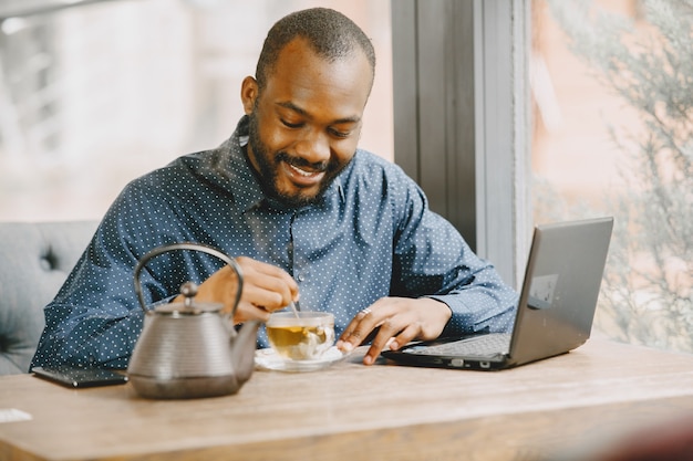 Afroamerikanischer Mann, der hinter einem Laptop arbeitet und in ein Notizbuch schreibt. Mann mit Bart sitzt in einem Café und trinkt einen Tee.