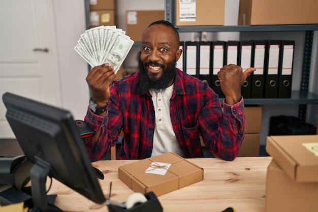 Afroamerikanischer Mann, der im E-Commerce von Kleinunternehmen arbeitet und Dollar hält, der mit dem Daumen nach oben zur Seite zeigt und glücklich mit offenem Mund lächelt