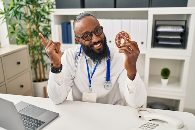 Afroamerikanischer Mann, der in einer Ernährungsklinik arbeitet und einen Donut in der Hand hält, lächelt glücklich und zeigt mit der Hand und dem Finger zur Seite