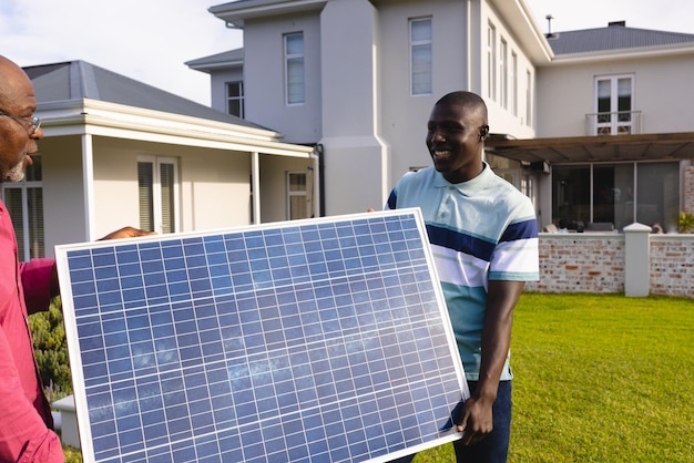 Afroamerikanischer Mann mit älterem Vater, der Solarpanel trägt, während er im Garten gegen ein Haus steht