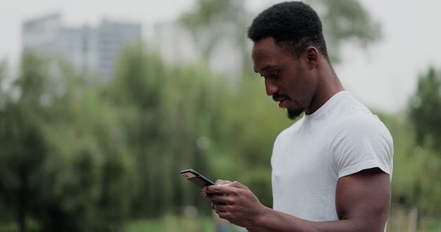 Afroamerikanischer Mann mit Smartphone im Stadtpark nach dem Training im Freien. Fitness Athlet Mann SMS mit Blick auf den Telefonbildschirm nach dem Training.