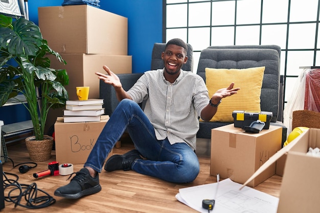 Afroamerikanischer Mann sitzt auf dem Boden im neuen Zuhause und lächelt fröhlich und bietet Hände an, die Hilfe und Akzeptanz geben.