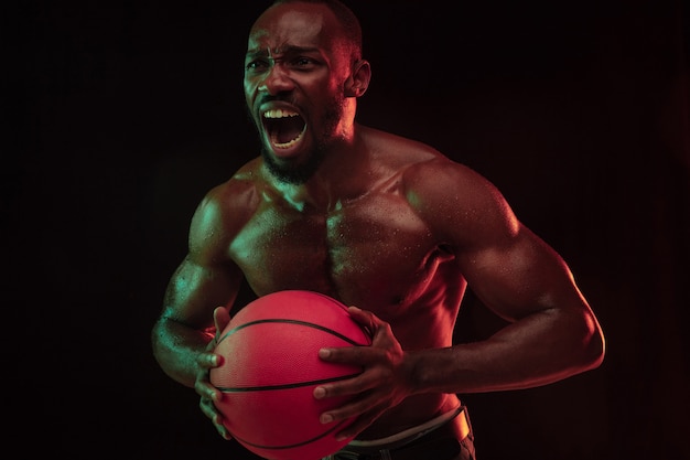 Afroamerikanischer muskulöser junger Basketballspieler in Aktion des Gameplay-Trainings, der in Neonlichtern über dunklem Studiohintergrund übt. Konzept von Sport, Bewegung, Energie, dynamischer, gesunder Lebensstil.