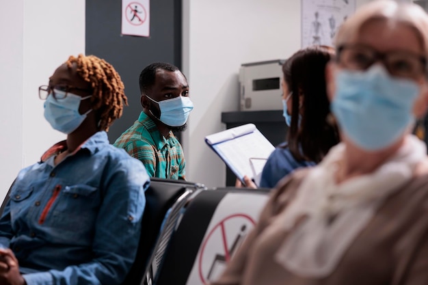 Afroamerikanischer Patient mit Gesichtsmaske, der mit der Krankenschwester spricht und im Wartebereich in der Empfangshalle des Krankenhauses sitzt. Mann, der Gesundheitsberatung mit medizinischem Personal in der Einrichtung durchführt.