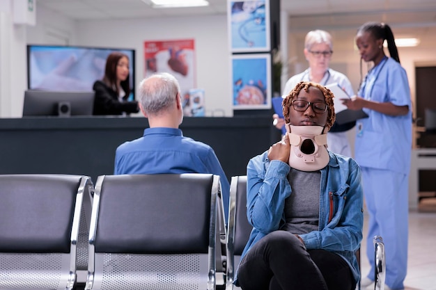 Afroamerikanischer Patient mit Halskragen, der im Wartezimmer des Krankenhauses sitzt, um an der Untersuchung teilzunehmen. Junger Erwachsener mit Verletzung, der einen Beratungstermin mit einem Spezialisten im Gesundheitszentrum hat.